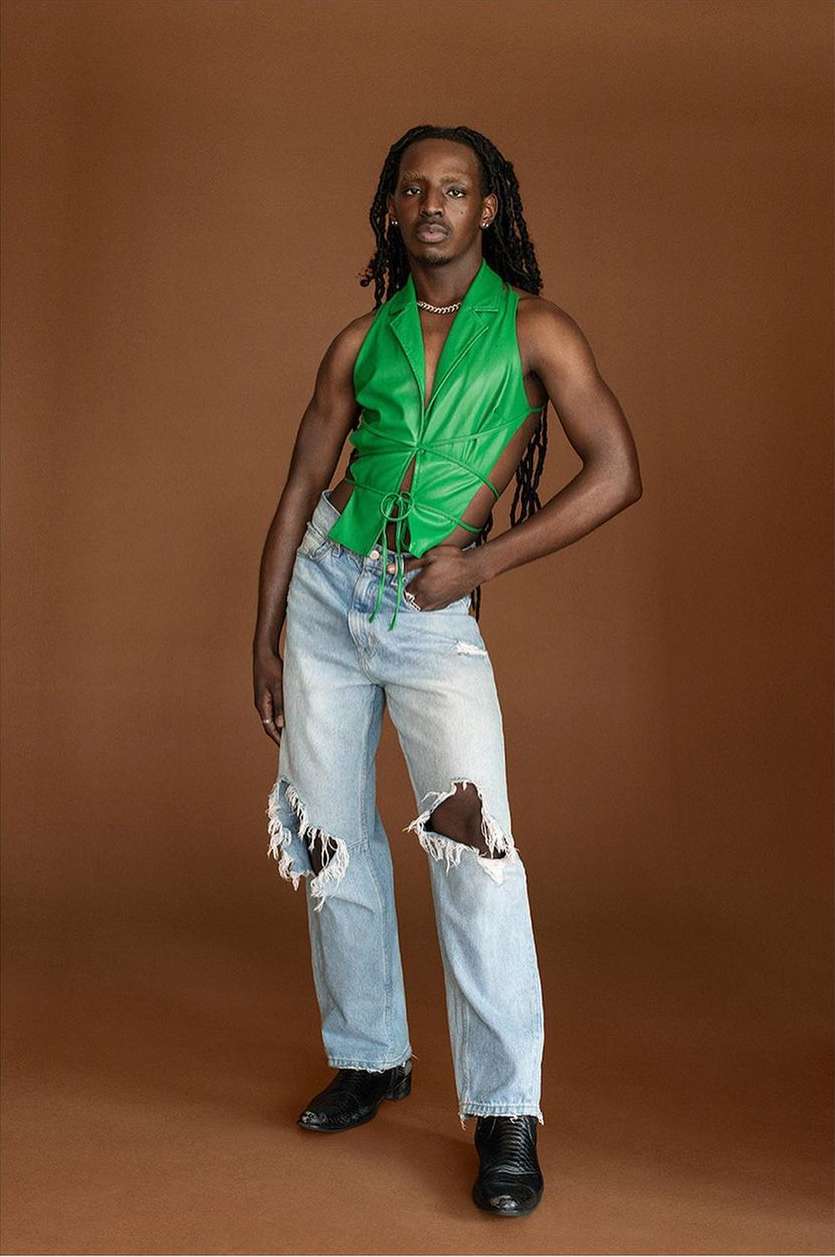 Black man with long dark dreadlocks wearing a bright green halter top and distressed light wash jeans against brown backdrop