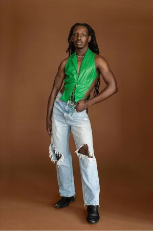 Black man with long dark dreadlocks wearing a bright green halter top and distressed light wash jeans against brown backdrop