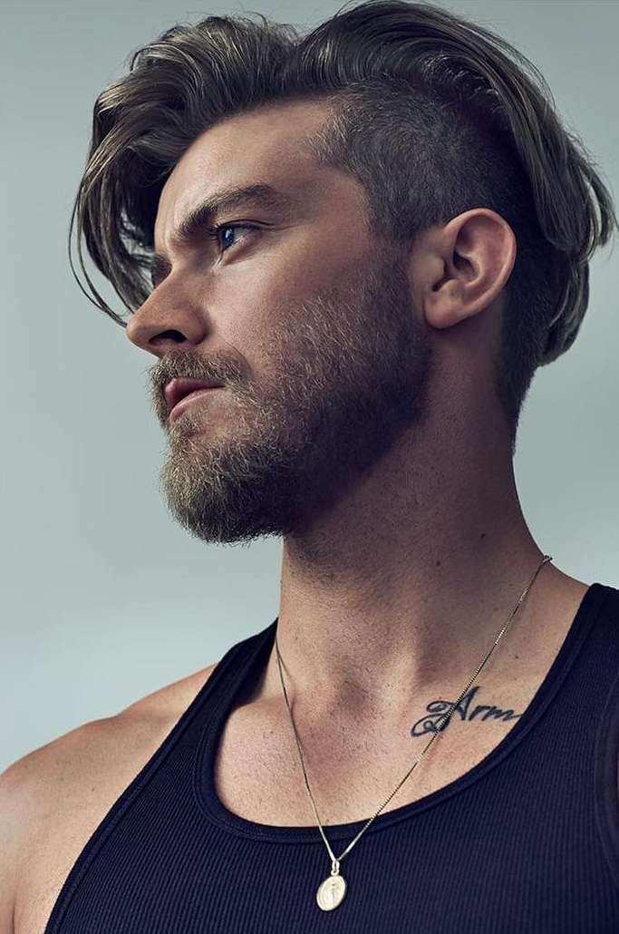 Studio shot of a bearded adult male with a shaved undercut and long hair, wearing a black tank top and gold necklace, looking to the side.