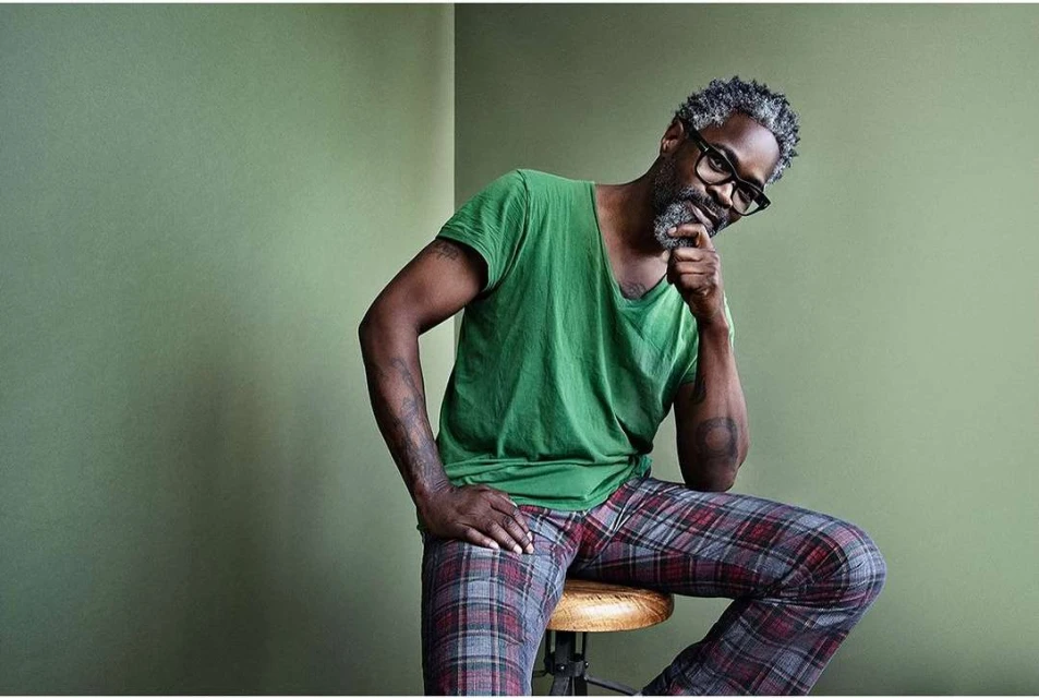 Adult male with grey beard and hair poses indoors, wearing a green t-shirt and plaid pajama pants, seated on a wooden stool.