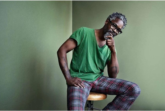 Adult male with grey beard and hair poses indoors, wearing a green t-shirt and plaid pajama pants, seated on a wooden stool.