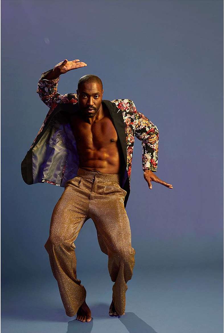 Shirtless Black male dancer poses in gold pants and floral jacket against a blue backdrop with arms up and legs in motion.