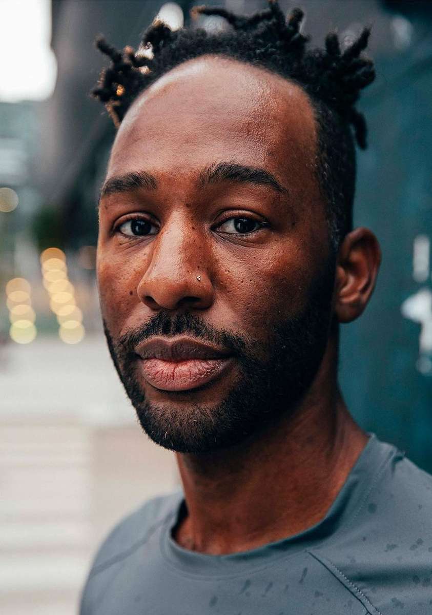 Portrait of handsome African American man with locs wearing grey top. Urban setting with blurred buildings and lights in the background.