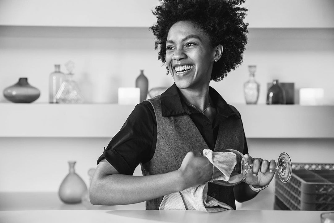 Monochrome portrait of a smiling African-American woman polishing a wine glass with a white cloth in an indoor setting, shelves in the background.