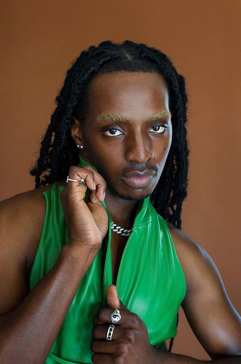 Portrait of a Black male model with dreadlocks wearing a green vest, silver chain and rings against a brown background in studio lighting.