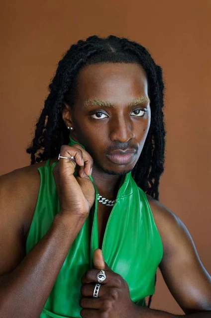 Portrait of a Black male model with dreadlocks wearing a green vest, silver chain and rings against a brown background in studio lighting.