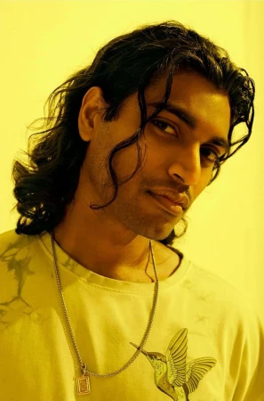 Close-up portrait of a man with long dark wavy hair wearing a graphic t-shirt and silver pendant necklace against a yellow backdrop.