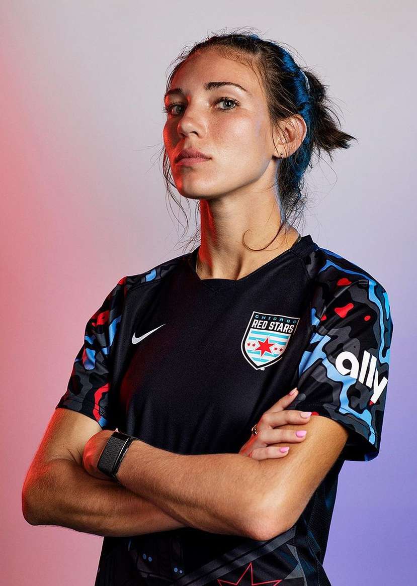 Portrait of a fair-skinned woman with arms crossed wearing a soccer jersey in front of a soft gradient background in a studio setting.