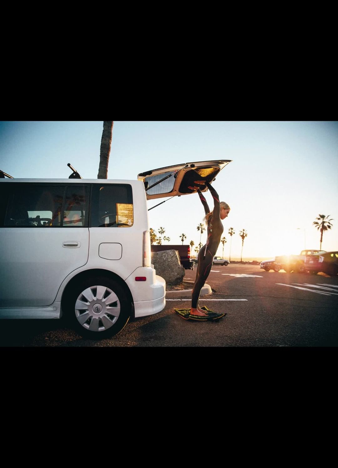 Woman in wetsuit loads surfboard into white SUV parked at beach near palm trees during golden hour, preparing for surfing.