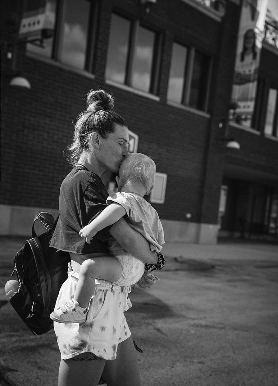 A mother kisses her child's head in a black and white photograph, while standing outdoors in an urban setting on a sunny day.