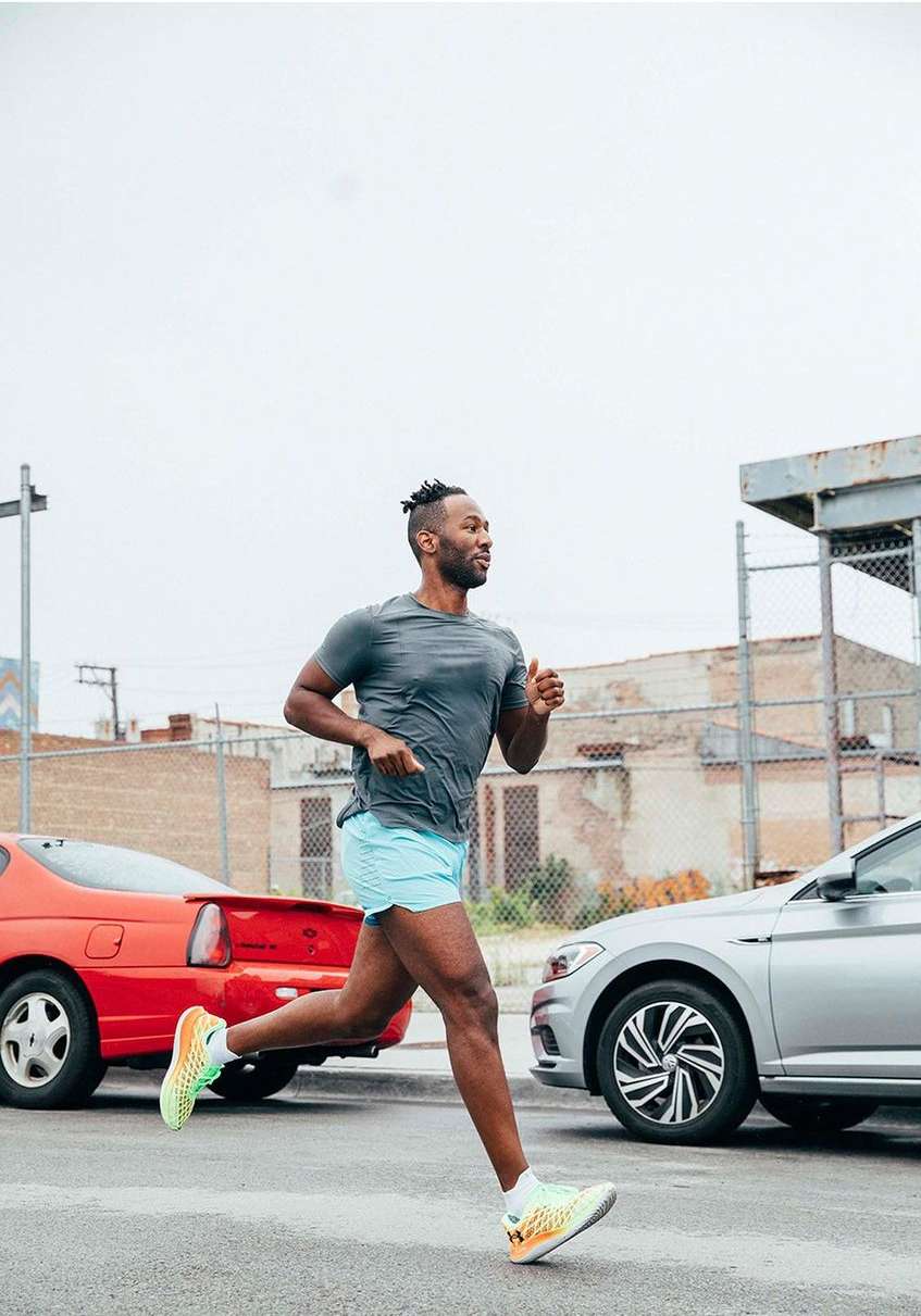 Athletic Black man running outdoors on city street wearing grey shirt and blue shorts with red and silver cars in the background.