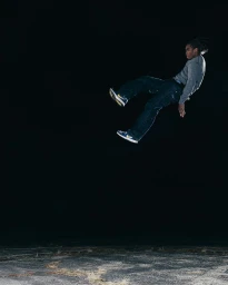 A Black man suspended mid-air in a dynamic jump against a dark background, wearing a grey top and blue jeans.