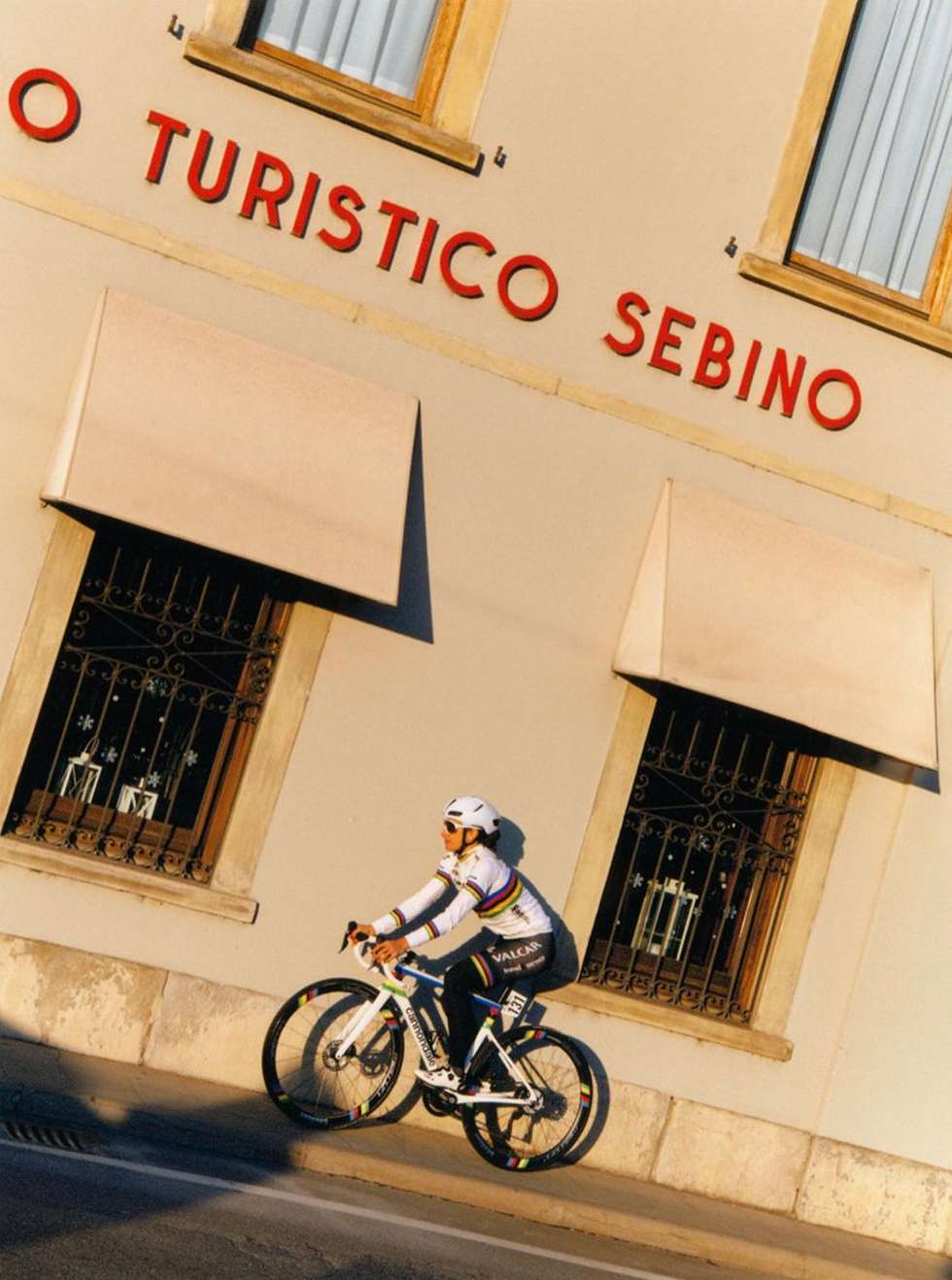 An cyclist rides a road bike near a building with awnings and signage in red lettering. The building casts a shadow on the road.