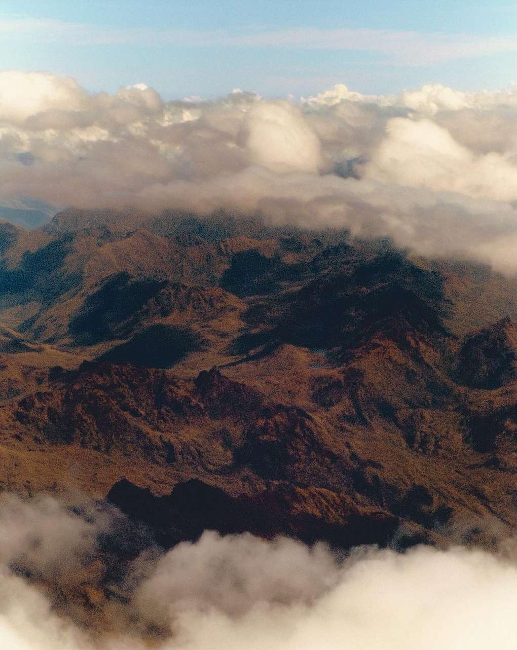 An aerial view of a mountainous landscape, partially obscured by clouds, bathed in soft daylight under a partly cloudy blue sky.