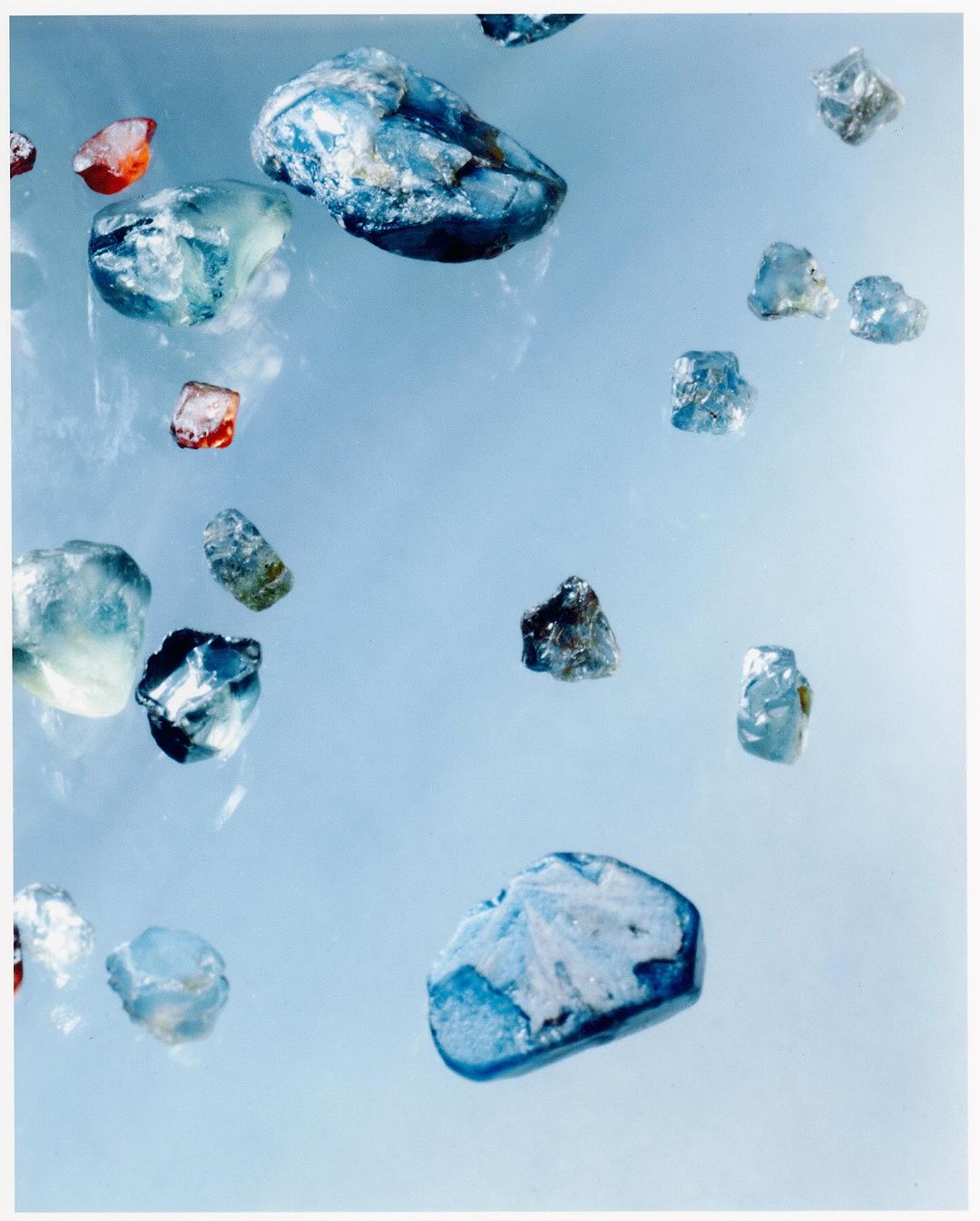 Scattered translucent gemstones with blue and red hues are displayed against a light blue background, suggesting a collection or raw materials.