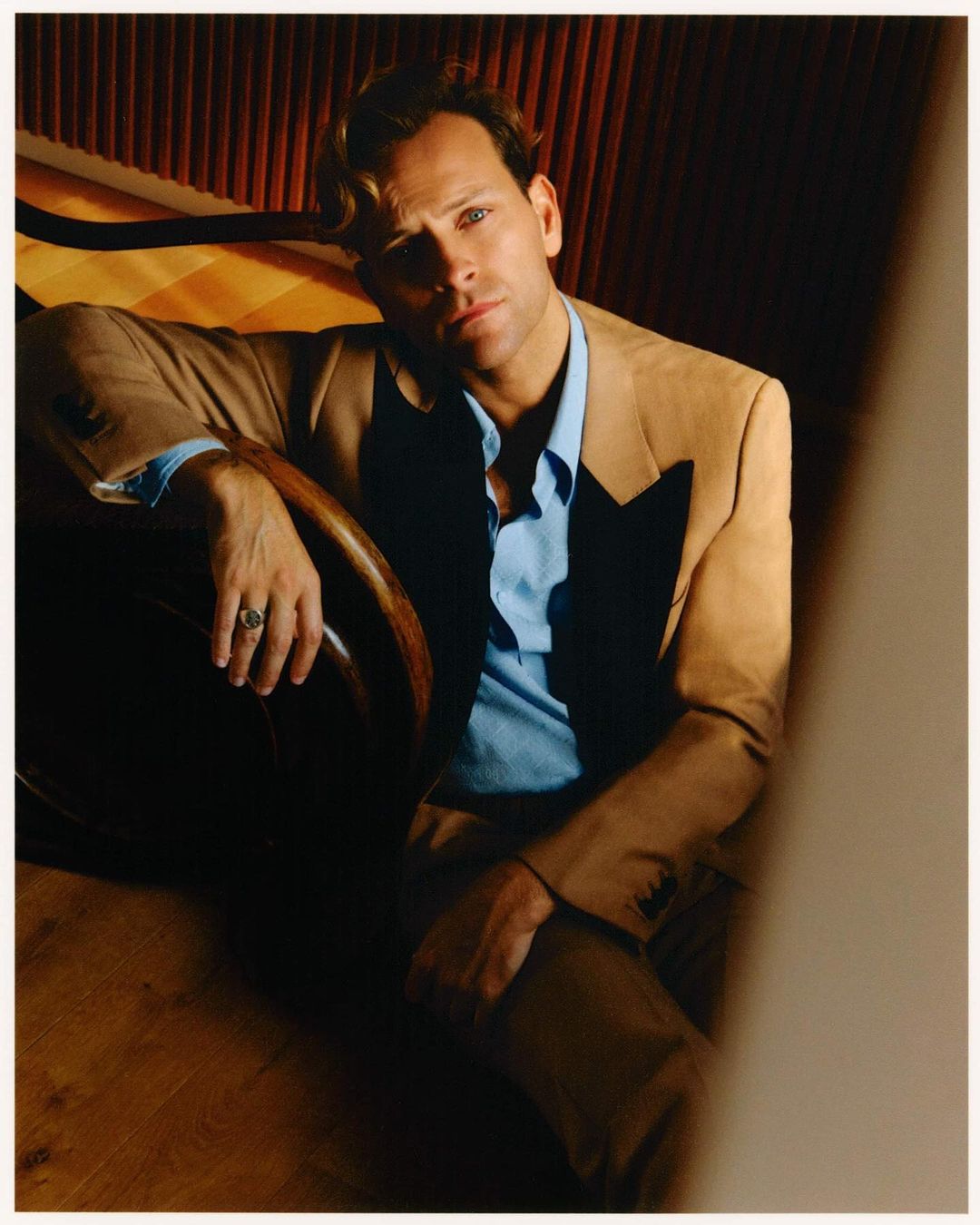 Portrait of a man in a beige suit and light blue shirt seated with an introspective expression, set against a dark, patterned backdrop.