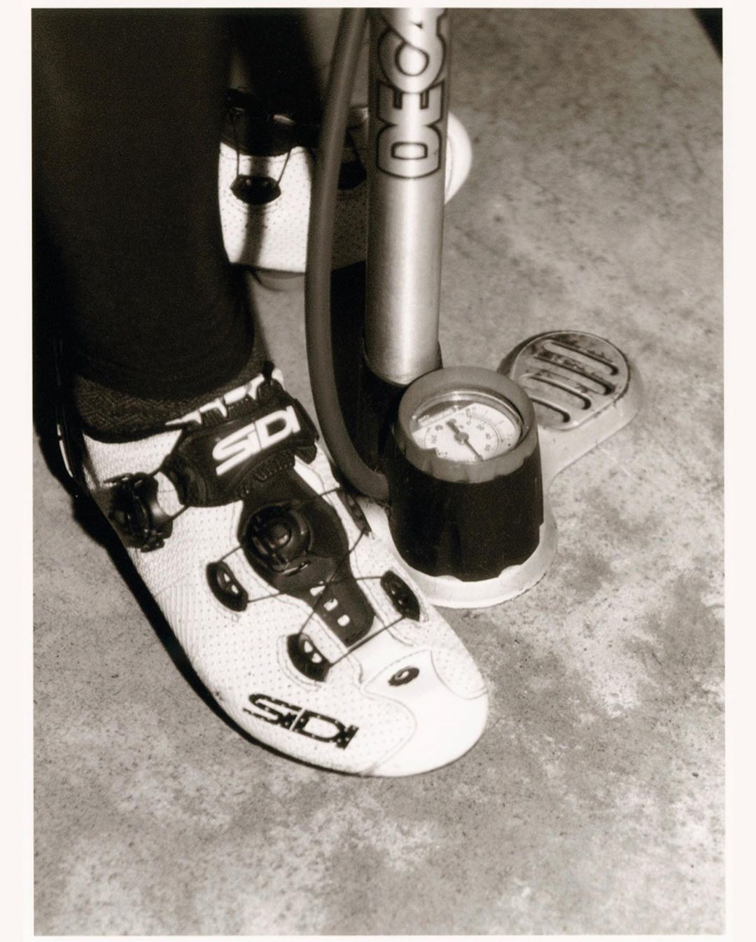 Close-up of a cyclist's foot in a white Sidi shoe attached to a bicycle pump on a gray concrete surface, low-angle view.