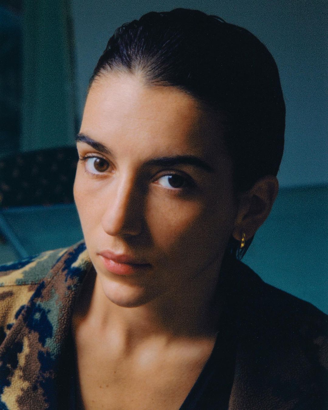 Close-up portrait of a young woman with dark hair and a camo jacket, looking directly at the camera, set against a dark background.