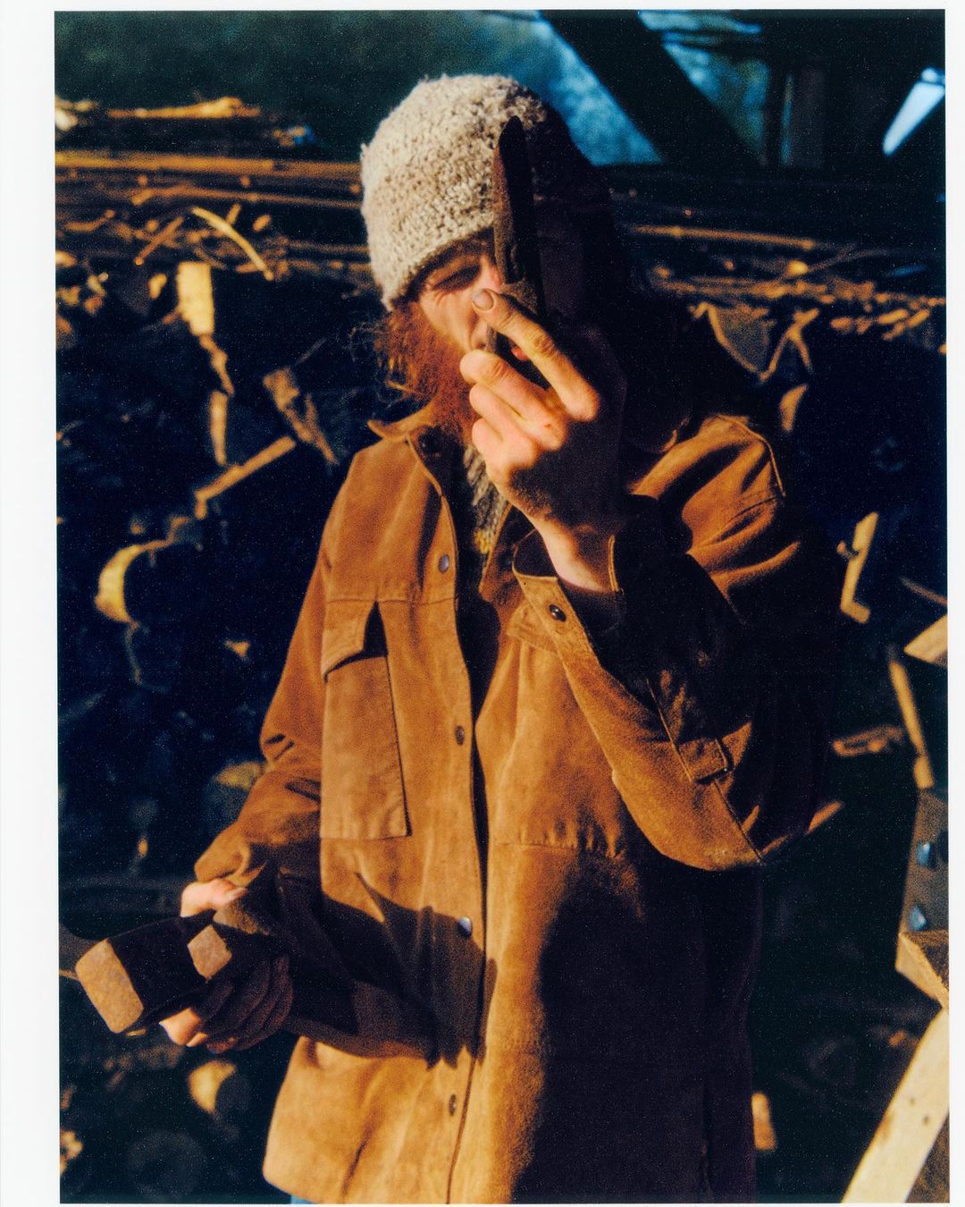 Adult with a red beard and grey knitted hat in a brown jacket holding a dark object, set against a background of wooden beams.