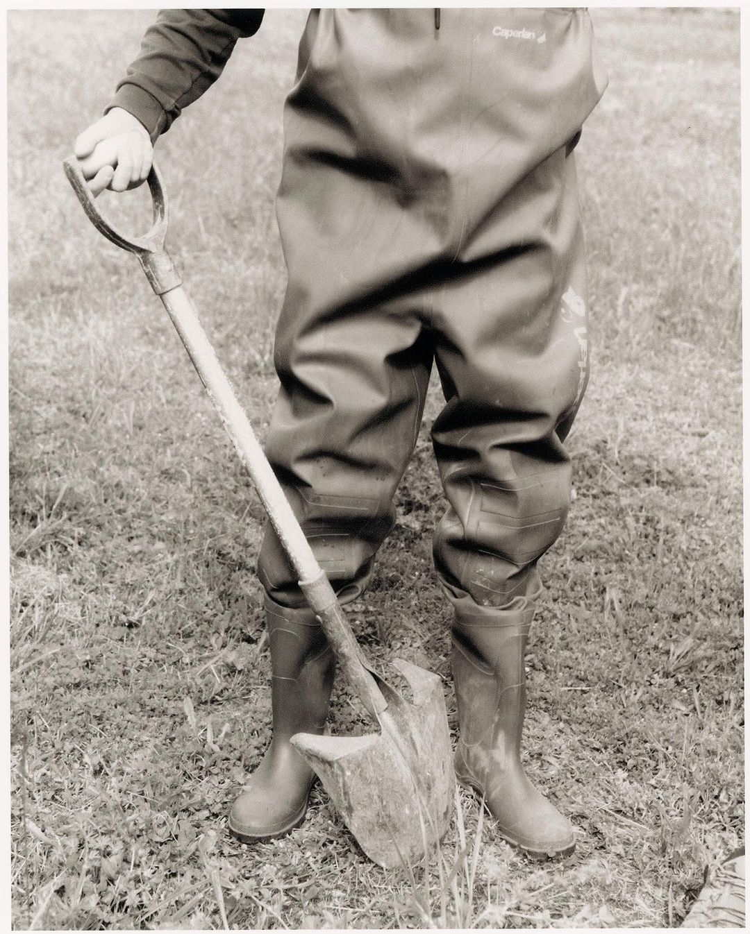 Person in waders stands outdoors holding a shovel, digging into the grass and dirt. The image is black and white with neutral tones.
