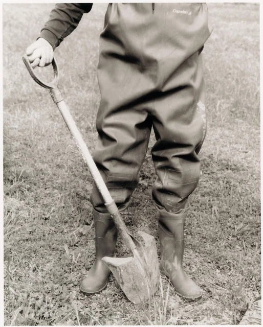 Person in waders stands outdoors holding a shovel, digging into the grass and dirt. The image is black and white with neutral tones.