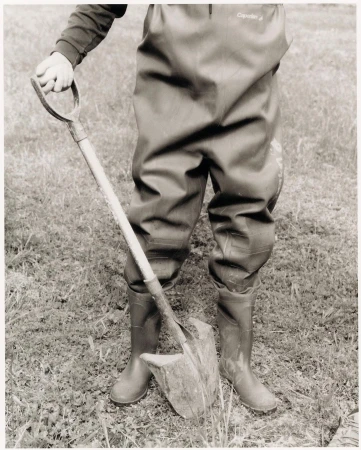 Person in waders stands outdoors holding a shovel, digging into the grass and dirt. The image is black and white with neutral tones.