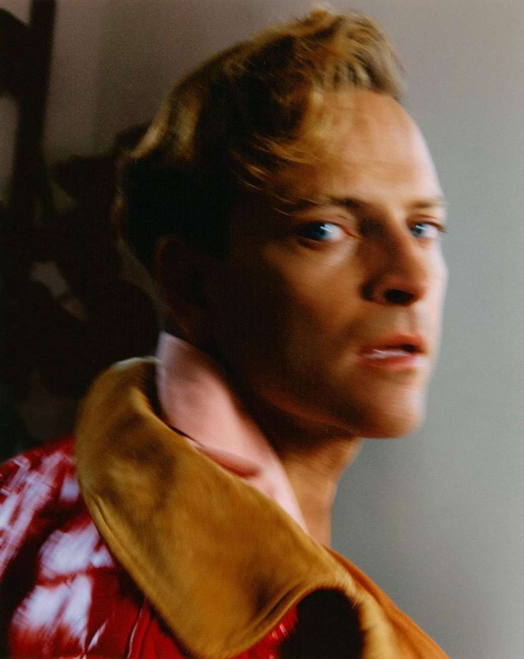 Close-up studio portrait of a fair-skinned adult male with blonde hair, wearing a red jacket with tan lapels and a pink collared shirt.