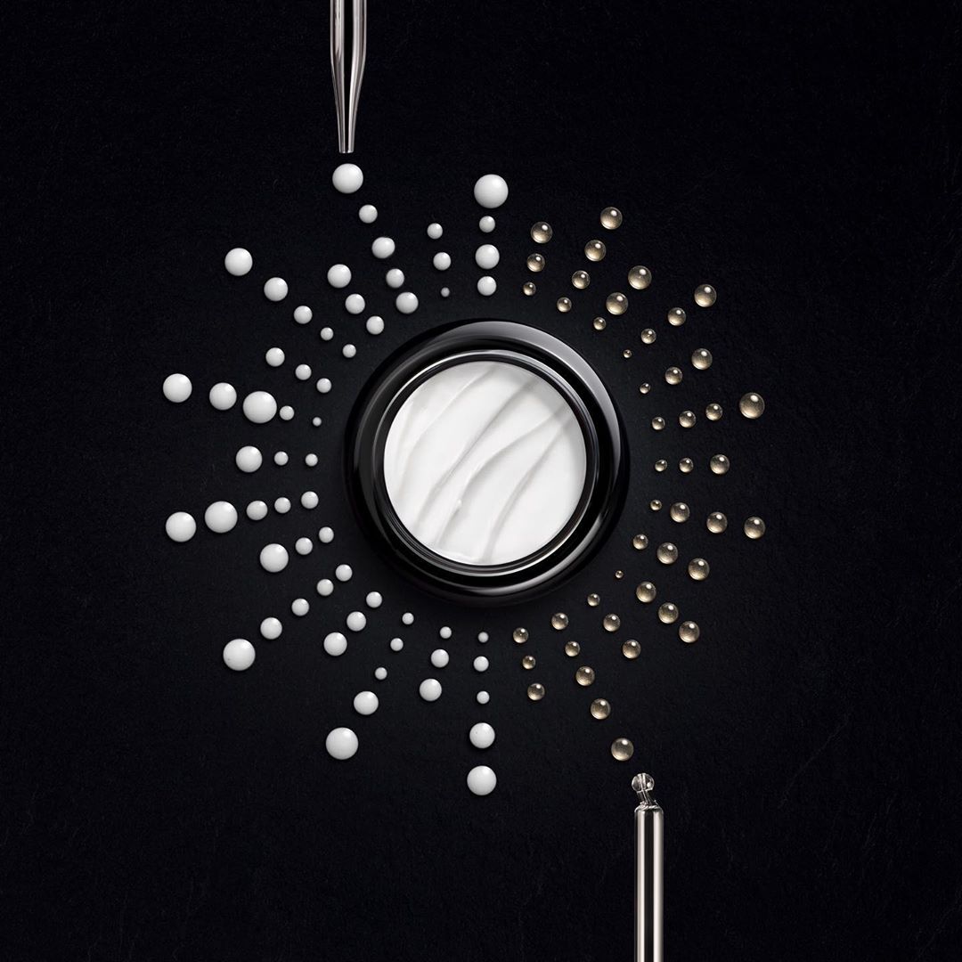 Close-up of a cream jar surrounded by white and gold liquid droplets, pipettes visible at top and bottom, on a black background.