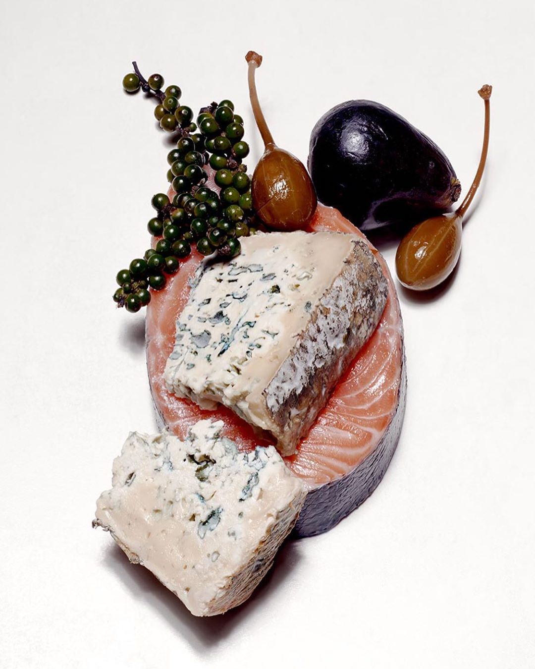 Overhead shot of salmon steak topped with blue cheese, green peppercorns, caper berries, and black olive on a white surface.