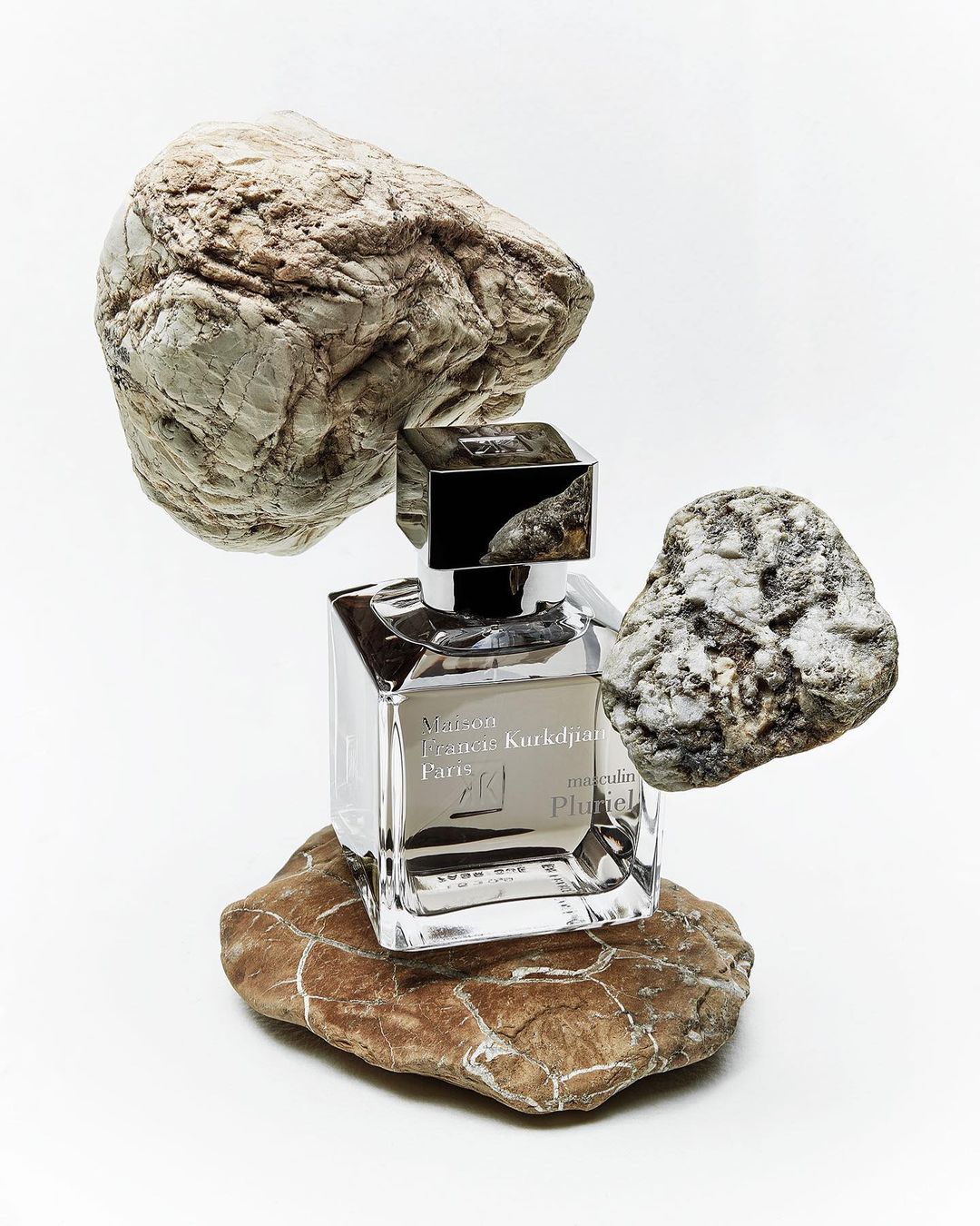 Maison Francis Kurkdjian Paris Pluriel perfume bottle elegantly balanced on textured rocks against a clean white backdrop.