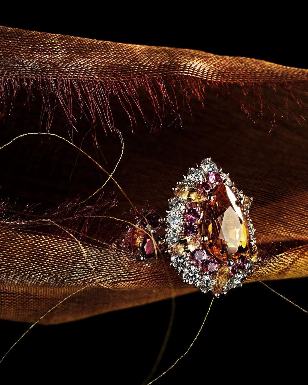 Close-up of an ornate gemstone ring with orange stone surrounded by smaller pink and white stones on brown textured fabric, on a black background.