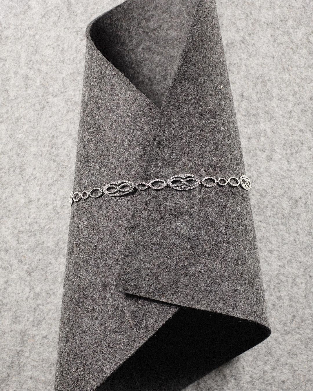 Silver chain with repeating infinity symbol links on folded grey felt fabric. Jewelry product shot showcases the necklace and material texture.