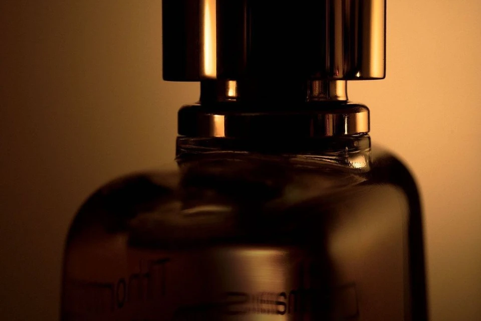 Close-up studio shot showcases the amber glass bottle of a perfume product with a gold cap against a blurred tan-colored backdrop.