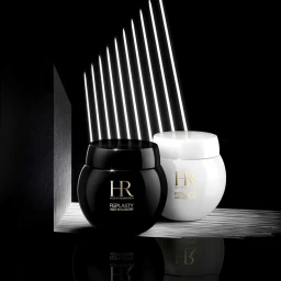 Two cosmetic jars, one black and one white, displayed on a textured surface beneath diagonal light beams against a black background.