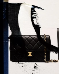 Black quilted leather handbag with gold chain strap positioned against a textured white background with bold black brush strokes.