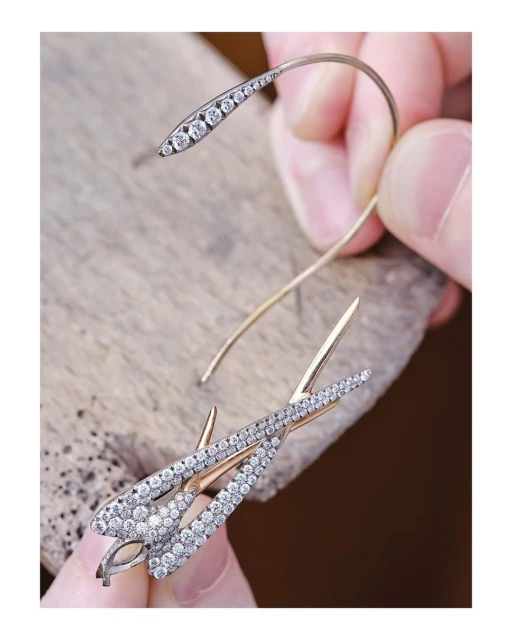 Close-up of a person's hands holding two handcrafted, gold, fish-shaped earrings covered in diamonds on a wooden surface.