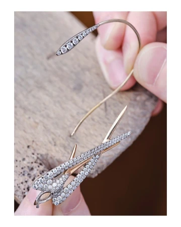 Close-up of a person's hands holding two handcrafted, gold, fish-shaped earrings covered in diamonds on a wooden surface.