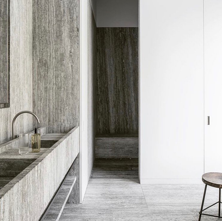 Modern minimalist bathroom interior with gray travertine stone walls, double sink, a steel faucet, toiletries, and a small stool.