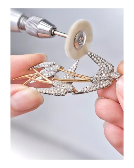 Close-up of two hands holding a diamond-studded gold swallow brooch, with a polishing tool touching the brooch in a light grey studio.