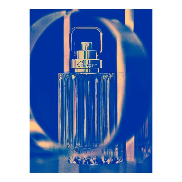 Close-up studio shot of Cartier Déclaration Haute Fraîcheur perfume bottle with clear glass and gold accents against a blurred blue background.