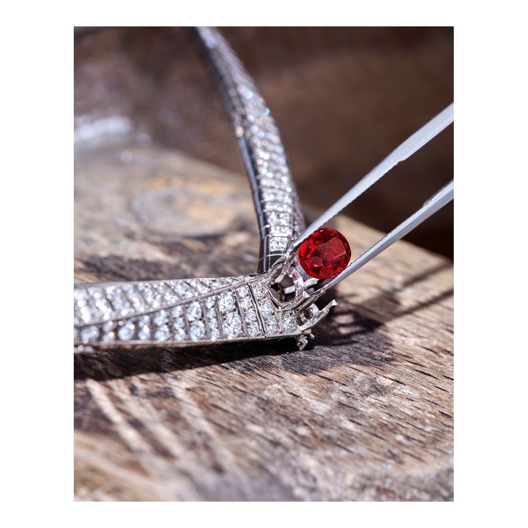 Close-up shot of a red gemstone being set in a diamond necklace with metal tweezers against a blurred wooden background. Jewelry setting.