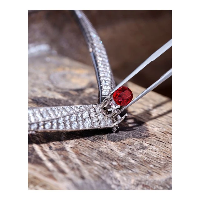 Close-up shot of a red gemstone being set in a diamond necklace with metal tweezers against a blurred wooden background. Jewelry setting.