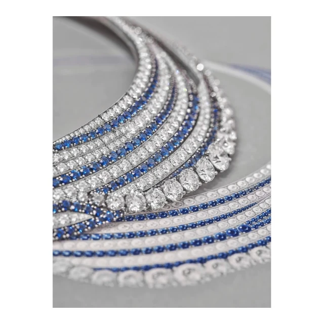 Close-up of a white gold necklace featuring diamonds and sapphires, laid flat against a neutral gray backdrop in a studio setting.