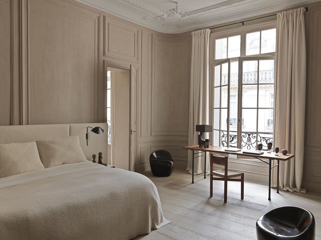 Interior of a bedroom with neutral tones, featuring a bed, desk with decor, chairs, and large windows with curtains, bathed in natural light.