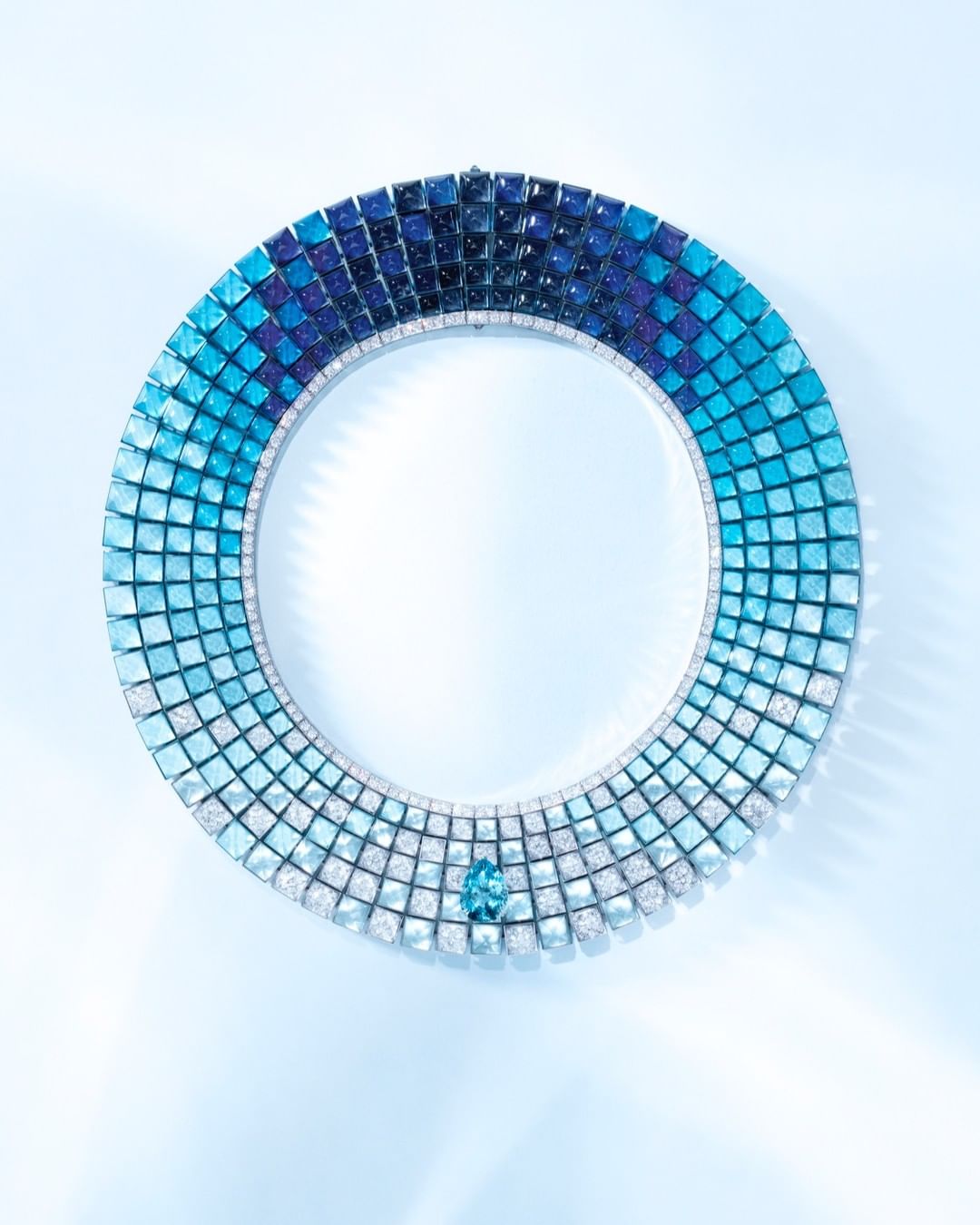 A circular necklace featuring a gradient of blue hues, adorned with a central blue gemstone, set against a soft blue backdrop.