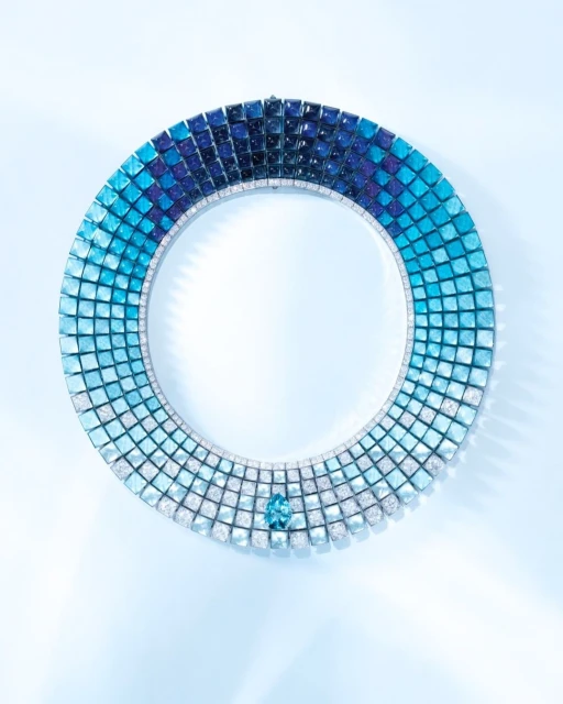 A circular necklace featuring a gradient of blue hues, adorned with a central blue gemstone, set against a soft blue backdrop.