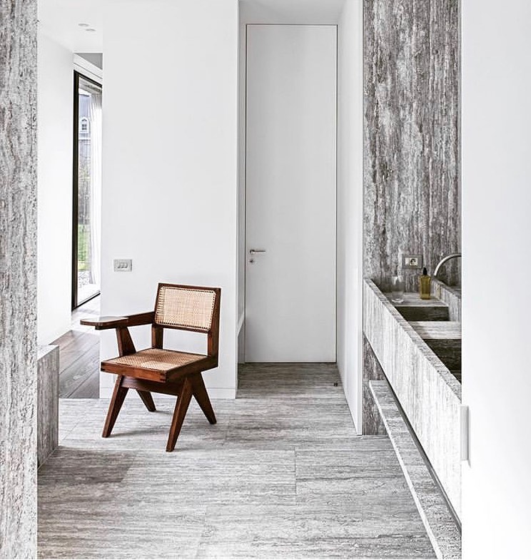 Minimalist interior of a home featuring a vintage wood and cane chair, gray stone walls, door, floor, and natural light.