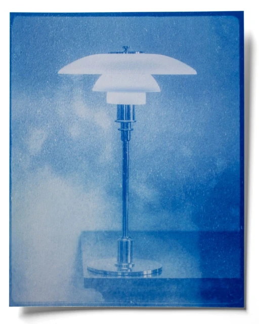 Minimalist cyanotype of a PH table lamp on a stand against a textured backdrop. The lamp has a white shade and chrome stem.