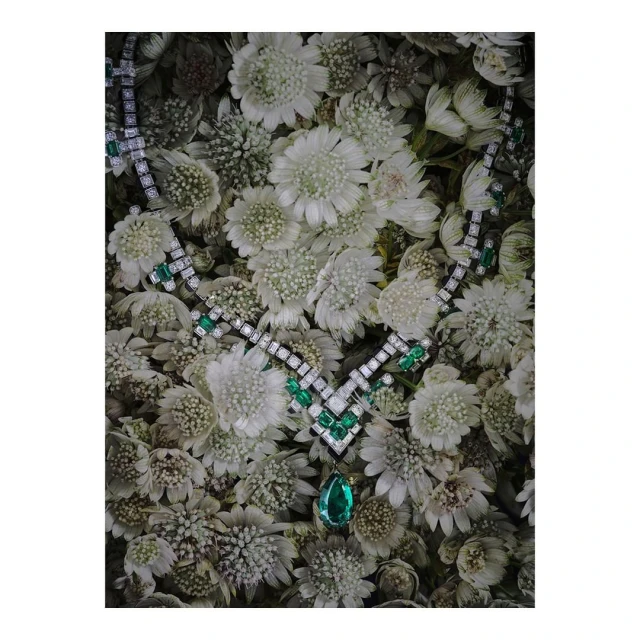 Elegant necklace with emerald and diamond gemstones displayed on a bed of white chrysanthemum flowers in a high-angle, still-life studio shot.