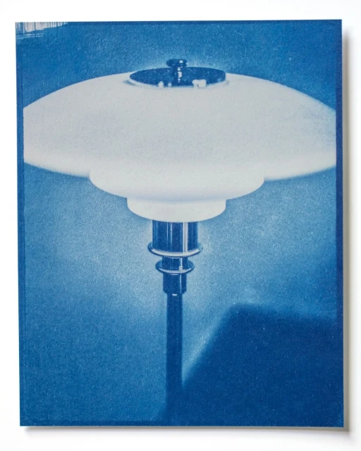 Cyanotype image of a vintage table lamp with a rounded white glass shade and a metal base sitting on a dark surface casting a shadow.